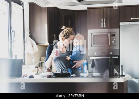 Glückliche Mutter hält ihren kleinen kleinen kleinen Jungen, während sie morgens Kaffee trinkt und Pfannkuchen zum Frühstück in der Hausküche macht. Familienleben, häusliches Lebenskonzept. Stockfoto