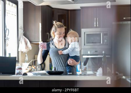 Glückliche Mutter hält ihren kleinen kleinen kleinen Jungen, während sie morgens Kaffee trinkt und Pfannkuchen zum Frühstück in der Hausküche macht. Familienleben, häusliches Lebenskonzept. Stockfoto