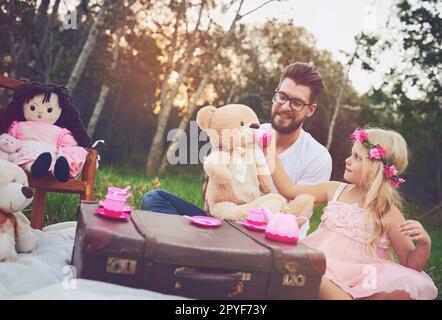 Hier ist ein Schluck für dich. Eine fröhliche Tochter und ein fröhlicher Vater, die eine Teeparty mit einem Haufen Stoffspielzeug mitten im Garten veranstalten. Stockfoto