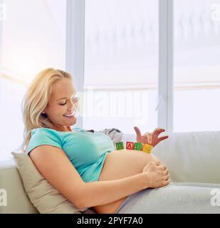 Schätze, Woll kommt bald. Eine schwangere Frau, die zu Hause mit Babyblöcken aus Holz auf dem Bauch liegt. Stockfoto