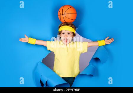 Basketballspiel. Überraschtes Kind mit Basketballball auf dem Kopf. Kleiner Basketballer in Sportbekleidung, der durch Papier schaut. Aktiver Sport. Der Junge spielt Stockfoto