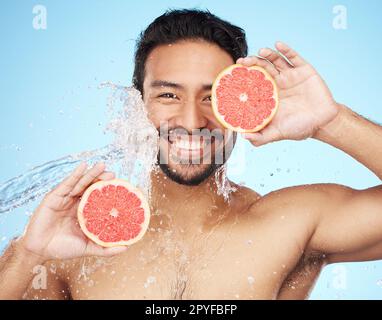 Gesicht, Wasser und Grapefruit mit einem männlichen Model im Studio auf blauem Hintergrund für Hygiene oder natürliche Flüssigkeitszufuhr. Hautpflege, Schönheit oder Obst mit einem gutaussehenden jungen Mann nass von einem Wasserspritzer in der Dusche Stockfoto