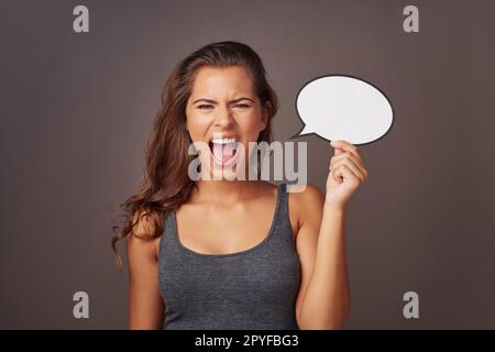 Sag es laut, sag es stolz. Studiofoto einer attraktiven jungen Frau, die eine leere Sprechblase hält und vor grauem Hintergrund schreit. Stockfoto