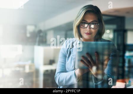 Shell hält ihre Termine mit den intelligentesten Apps ein. Eine junge Geschäftsfrau, die lange auf einem digitalen Tablet in einem Büro arbeitet. Stockfoto