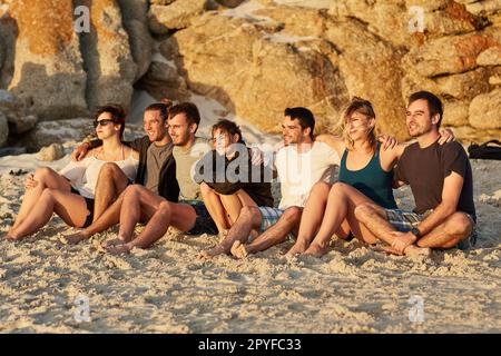 Einen Sonnenuntergang mit Freunden teilen. Eine Gruppe glücklicher junger Freunde, die zusammen den Sonnenuntergang bewundern, während sie am Strand sitzen. Stockfoto