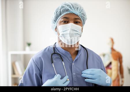 Portrait des afroamerikanischen männlichen in blauen Peelings tragen Gesichtsmaske, medizinische Kappe und Latexhandschuhe halten Stethoskop um den Hals am Arbeitsplatz. Gesundheitsexperte bereitet sich auf die Untersuchung vor. Stockfoto