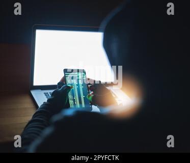 In der schattigen Welt des Online-Hacking. Ein nicht erkennbarer Hacker, der im Dunkeln ein Handy und einen Laptop benutzt. Stockfoto