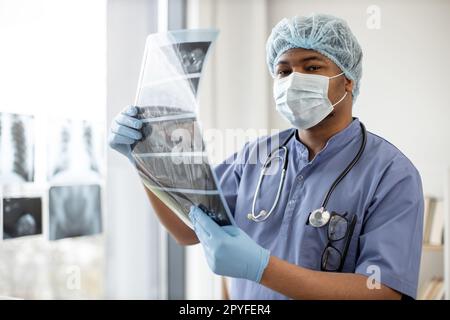 Aufmerksamer multiethnischer Mann in Schutzkleidung hält detailliertes CT-Scan-Bild des Körperteils während des Aufenthalts im Untersuchungsraum des Krankenhauses. Arztpraxis Stockfoto