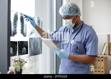 Aufmerksamer multiethnischer Mann in medizinischen Peelings und Gesichtsmaske, der Röntgenbilder betrachtet, während er Tabletten am Arbeitsplatz des Arztes hält. Arzt in Handschuhen und Kappenuntersuchung von Thoraxaufnahmen im Krankenhausinnenraum. Stockfoto