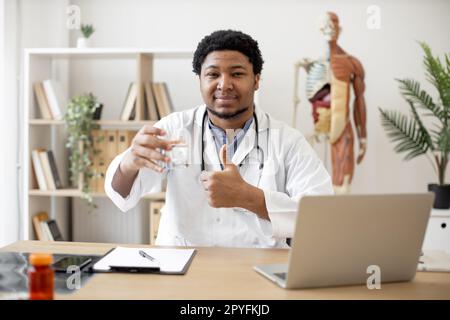 Selbstbewusster multiethnischer Therapeut am Schreibtisch, der halb volles Glas hält und ein Daumenschild zeigt, während er im Sprechzimmer arbeitet. Hausarzt genehmigt Vorteile von sauberem Wasser für das Gesundheitswesen. Stockfoto