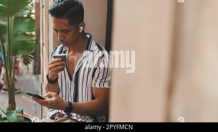 Der junge Mann sitzt draußen und trinkt Kaffee und benutzt ein Handy Stockfoto