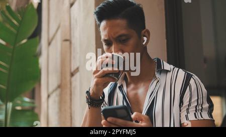 Der junge Mann sitzt draußen und trinkt Kaffee und benutzt ein Handy Stockfoto