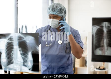 Durchdachter, multiethnischer Erwachsener in blauen Peelings und Gesichtsmaske, der am Telefon spricht, während er Röntgenbilder in einer Praxispraxis scannt. Therapeut diskutiert Gesundheitszustand mit Patient. Stockfoto