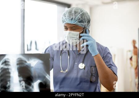 Durchdachter, multiethnischer Erwachsener in blauen Peelings und Gesichtsmaske, der am Telefon spricht, während er Röntgenbilder in einer Praxispraxis scannt. Therapeut diskutiert Gesundheitszustand mit Patient. Stockfoto