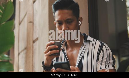 Der junge Mann sitzt draußen und trinkt Kaffee und benutzt ein Handy Stockfoto