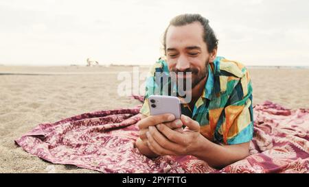 Ein lächelnder Mann liegt am Strand mit Smartphone in der Hand Stockfoto