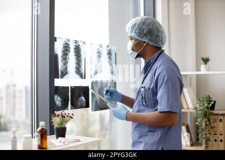 Aufmerksamer multiethnischer Mann in medizinischen Peelings und Gesichtsmaske, der Röntgenbilder betrachtet, während er Tabletten am Arbeitsplatz des Arztes hält. Arzt in Handschuhen und Kappenuntersuchung von Thoraxaufnahmen im Krankenhausinnenraum. Stockfoto