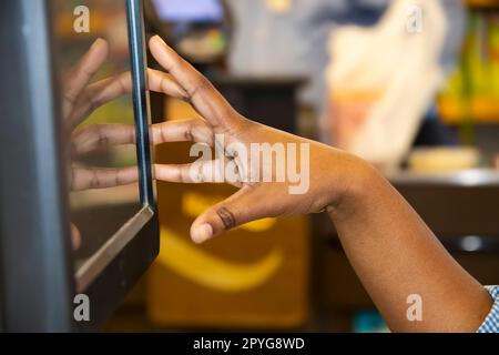 Die Hand einer schwarzen Kassiererin mit Touchscreen-Kasse und verschwommenem Bokeh-Hintergrund – Reflexion der Hand auf dem Bildschirm Stockfoto