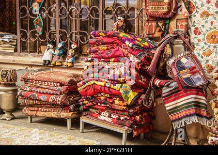 Stapel von Teppichen und mit Suzani bestickten und dekorativen Tribal-Textilien - und Taschen, Puppen und andere Gegenstände, die außerhalb des Tiflis Georgia Store zum Verkauf gestapelt werden Stockfoto