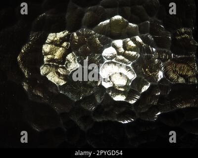 Wellglas, Rücklichtquelle. Spiel von Blendungen und Reflexionen auf einer transparenten, unebenen Oberfläche. Dunkle und helle abstrakte asymmetrische, mehrfach beleuchtete Ebenen. Abstrakter, holpriger goldener Hintergrund. Stockfoto