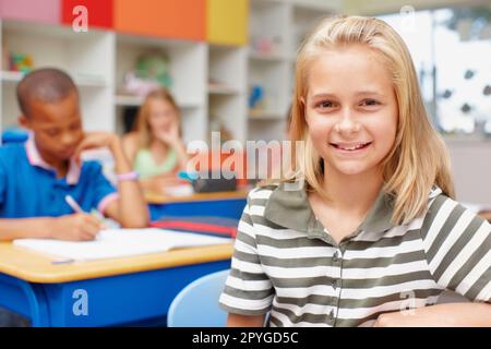 Sie ist stolz auf ihre Schule und ihre Bildung. Selbstbewusstes, junges Schulmädchen, das im Unterricht an ihrem Schreibtisch sitzt und die Kamera anlächelt - Kopierraum. Stockfoto