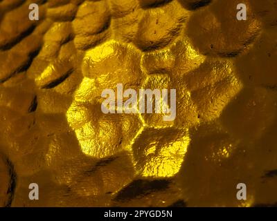 goldgelbes Wellglas mit hellem und dunklem Hintergrund. Lichtreflexionen. Wellglas, Rücklichtquelle. Spiel von Blendungen und Reflexionen auf einer transparenten, unebenen Oberfläche. Stockfoto