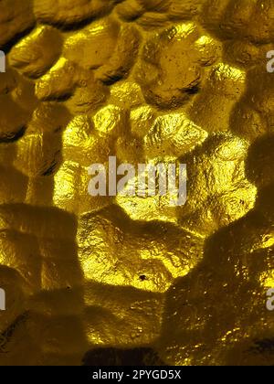goldgelbes Wellglas mit hellem und dunklem Hintergrund. Lichtreflexionen. Wellglas, Rücklichtquelle. Spiel von Blendungen und Reflexionen auf einer transparenten, unebenen Oberfläche. Stockfoto