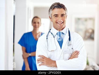 Ich konnte es nicht ohne ihre Unterstützung tun. Porträt eines männlichen Arztes, der im Krankenhaus mit gekreuzten Armen steht, mit einer weiblichen Kollegin im Hintergrund. Stockfoto