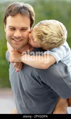 Die Liebe zu einem Sohn. Der Junge küsst seinem Vater die Wange, während er ihm Huckepack gibt. Stockfoto