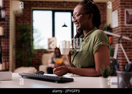 Lächelnder Programmierer, der eine Pause von der Arbeit an einem entfernten Ort macht und eine Tasse Kaffee oder Tee im Wohnzimmer hält. Freiberufler, der sich entspannt und Videoinhalte auf dem Computerbildschirm ansieht, während ein Mitbewohner liest. Stockfoto