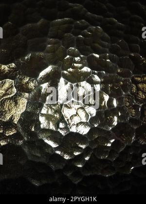 Wellglas, Rücklichtquelle. Spiel von Blendungen und Reflexionen auf einer transparenten, unebenen Oberfläche. Dunkle und helle abstrakte asymmetrische, mehrfach beleuchtete Ebenen. Abstrakter, holpriger goldener Hintergrund. Stockfoto