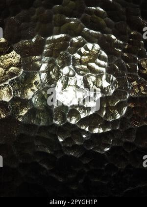 Wellglas, Rücklichtquelle. Spiel von Blendungen und Reflexionen auf einer transparenten, unebenen Oberfläche. Dunkle und helle abstrakte asymmetrische, mehrfach beleuchtete Ebenen. Abstrakter, holpriger goldener Hintergrund. Stockfoto