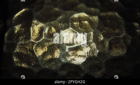 Wellglas, Rücklichtquelle. Spiel von Blendungen und Reflexionen auf einer transparenten, unebenen Oberfläche. Dunkle und helle abstrakte asymmetrische, mehrfach beleuchtete Ebenen. Abstrakter, holpriger goldener Hintergrund. Stockfoto