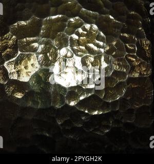 Wellglas, Rücklichtquelle. Spiel von Blendungen und Reflexionen auf einer transparenten, unebenen Oberfläche. Dunkle und helle abstrakte asymmetrische, mehrfach beleuchtete Ebenen. Abstrakter, holpriger goldener Hintergrund. Stockfoto