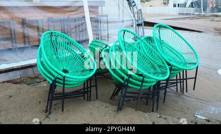 Korbgrüne Plastikstühle mit schwarzen Beinen in einem Café an einem Sandstrand nach einem Sturm. Die Möbel sind übereinander gestapelt, da das Strandcafe geschlossen ist. Ende der Strandsaison im Hotel Stockfoto