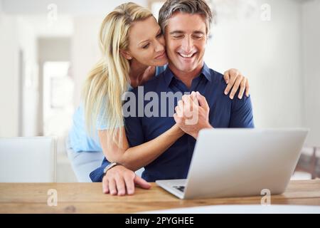 Sie hilft mir bei jedem Schritt. Eine attraktive, reife Frau, die ihren hübschen, reifen Ehemann umarmt, während er ein Notebook im Haus benutzt. Stockfoto