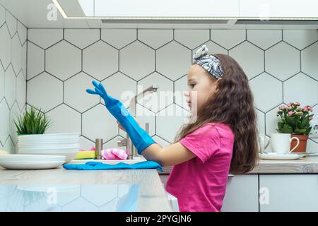 Nettes kleines Mädchen, das blaue Haushaltshandschuhe anzieht, um Geschirr abzuspülen. Hausreinigung, persönliche Schutzausrüstung. Stockfoto