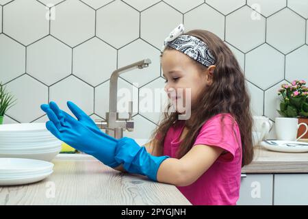 Glückliches kleines Mädchen, das blaue Haushaltshandschuhe anzieht, um Geschirr in der Küchenspüle zu spülen. Hausreinigungskonzept. Stockfoto