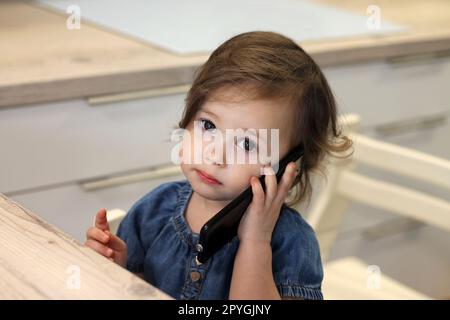 Süßes, kleines, dunkelhaariges Mädchen, 1,5-2,5, in einem Jeanskleid, spricht mit jemandem über ein Smartphone in der Küche. Stockfoto