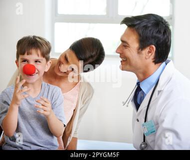 Das erste Anzeichen einer Grippe ist eine rote Nase. Ein süßer kleiner Junge, der mit einer Clownsnase im Arztzimmer spielt. Stockfoto