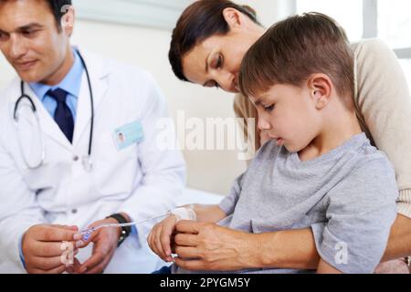 Der Arzt wird sich sehr gut um Sie kümmern. Ein kleiner Junge, der auf seine Infusion schaut, während seine Mutter ihn festhält. Stockfoto