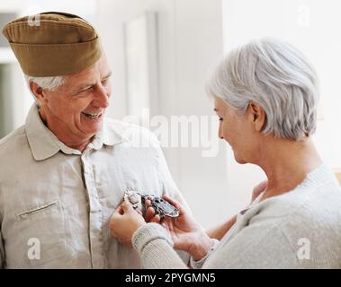 Ich bin so stolz auf meinen Kriegsveteranen-Ehemann. Eine ältere Frau, die sich die Medaillen ihres Mannes ansieht, während er steht und seine komplette Armeeuniform trägt. Stockfoto