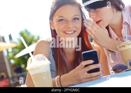 Verbreiten Sie es... Ein Teenager, das SMS auf einem Handy schreibt, während ihr Freund ihr etwas ins Ohr flüstert. Stockfoto