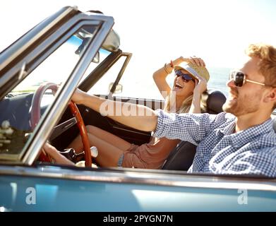 Halt dich an deinem Hut fest, Schatz. Ein glückliches junges Paar, das an einem schönen Sommertag in einem Cabrio fährt. Stockfoto