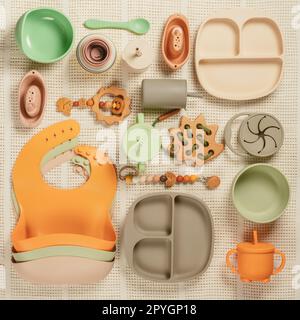 Pastellfarbenes Silikonset mit Geschirr, Besteck, Lätzchen, Accessoires und Holzspielzeug für Kinder auf weißem Stoffhintergrund. Stockfoto