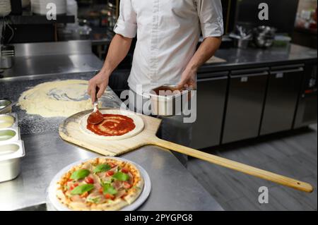 Männlicher Koch, der Tomatenpaste auf Pizzabasis ausstreut Stockfoto