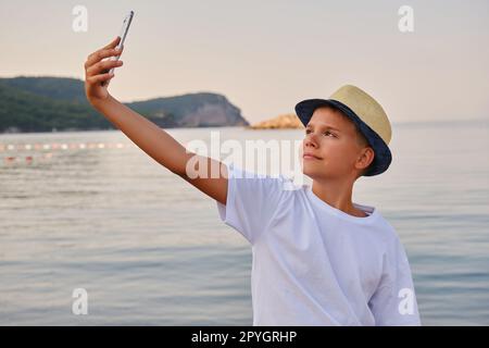 Ein Junge mit Hut macht ein Selfie-Porträt am Meer. Glücklicher Kerl, der Sommerferien am Strand genießt Stockfoto