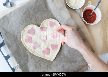 Herzförmiger Pizzateig, der Koch legt herzförmige Wurst. Italienische Pizza, herzförmige Pizza. Stockfoto