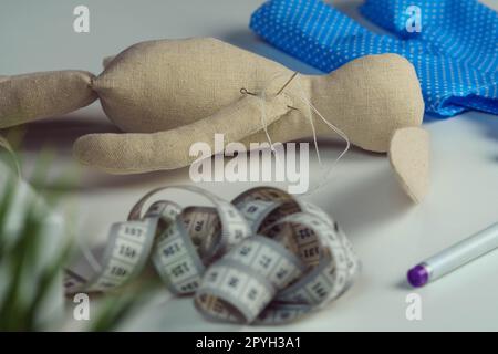 Nähen von gefüllten Kaninchenspielzeugen mit Nadel im Körper, die auf dem Tisch liegt. Strick Hobby. Hase in der Nähe von blauem Textil mit Punktmuster Stockfoto