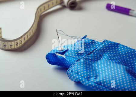 Kleines niedliches blaues, gepunktetes Kostüm mit Nadelanzug für Spielzeug in der Nähe von Maßband und Stift auf weißem Tisch. Nähen Stockfoto